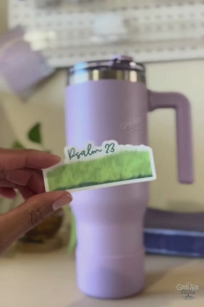  vertical video clip of a small business owner peeling a high-quality waterproof vinyl sticker that features green text reading "Psalm 23" over a hand-drawn green grass graphic. The sticker is smoothly applied to a solid purple water bottle.
