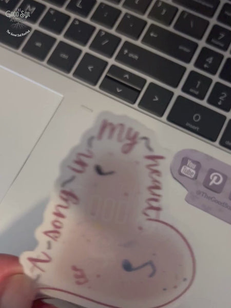Hand placing clear vinyl sticker onto the palm area of a laptop. Sticker features a heart design with music notes.